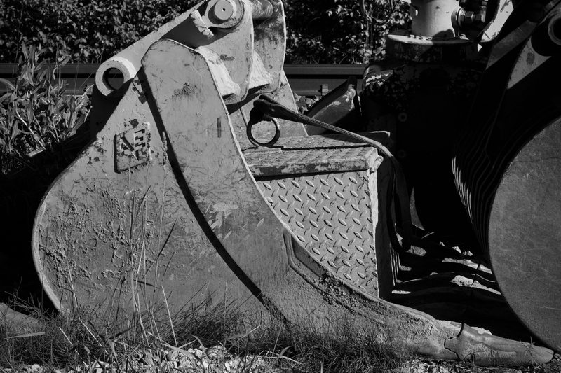 Black and white close-up of the steel bucket of an excavator, with a tread-plate panel and bolted joints beside it, on grass.